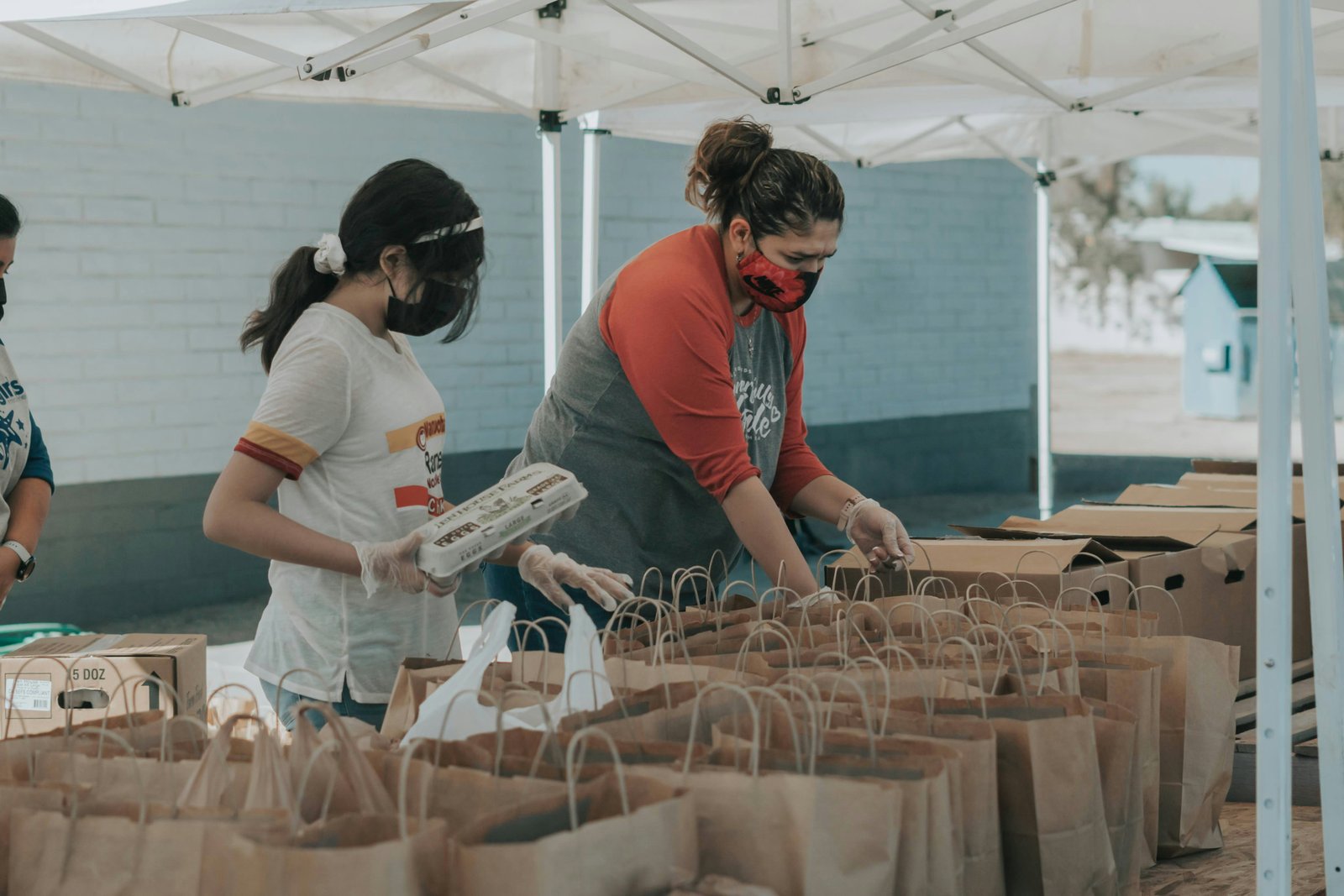Volunteers packing paper bags with food and supplies under a tent.