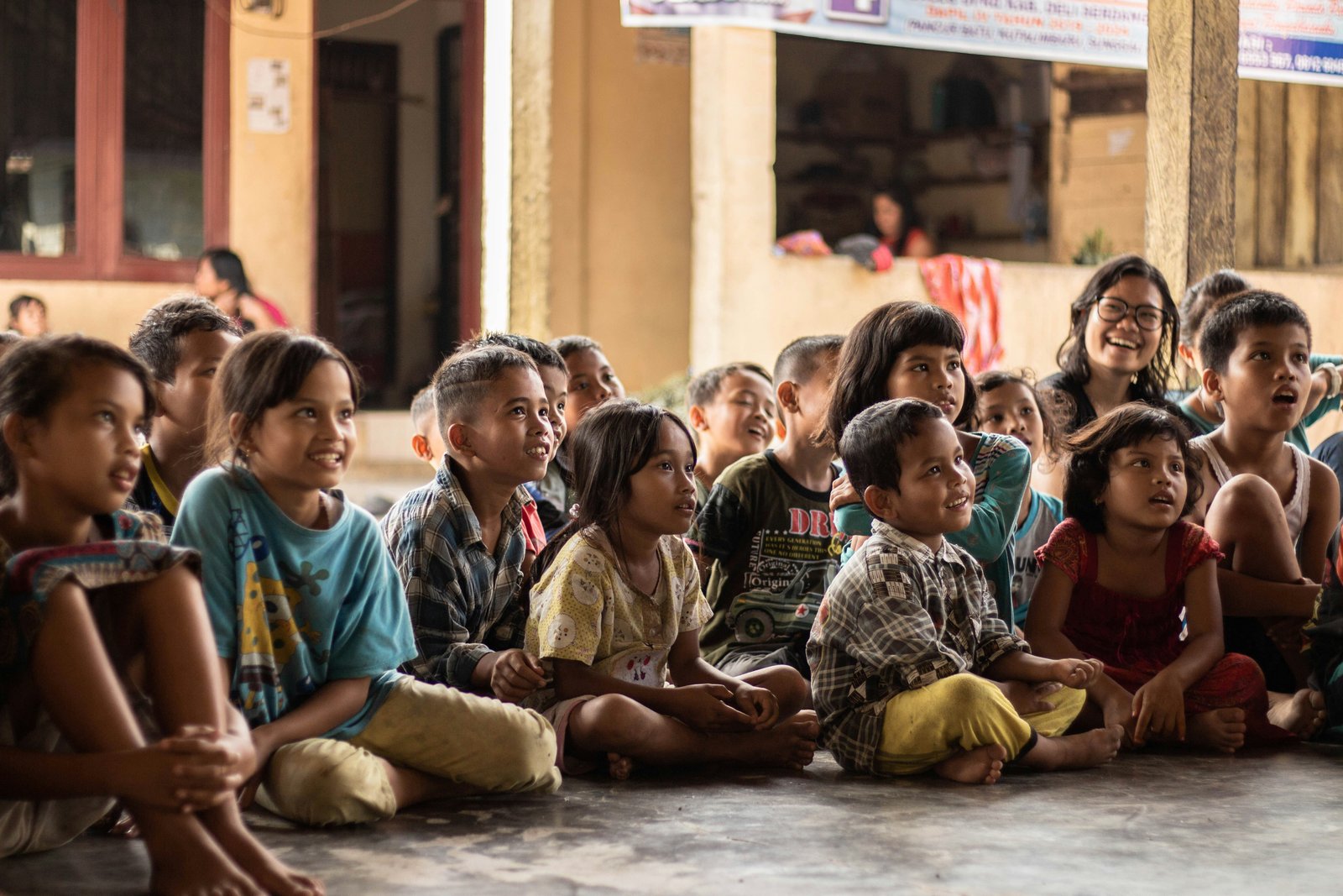 Children gathered outdoors, smiling and listening as a volunteer teaches a health lesson.