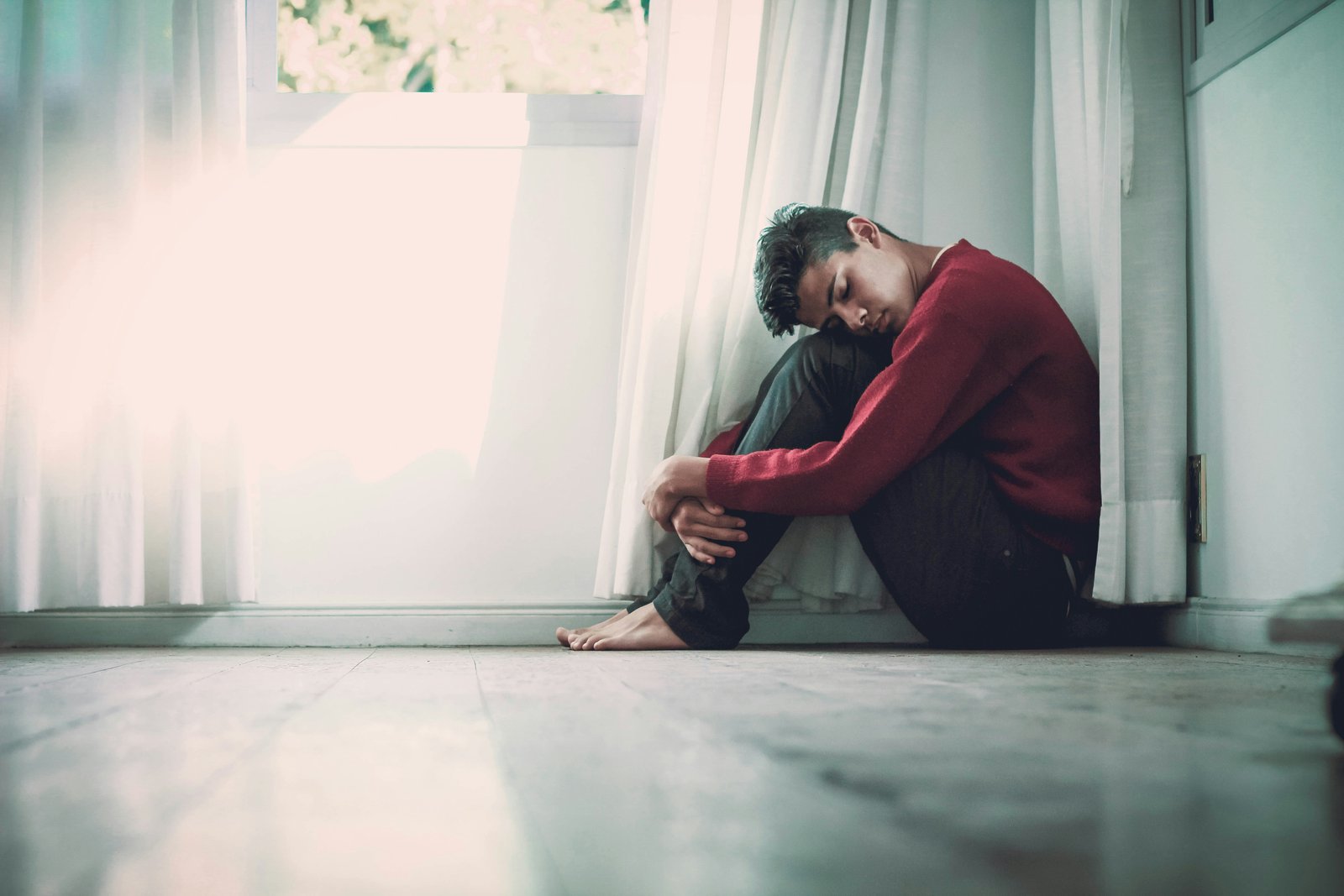 A young man sitting alone on the floor by a window, head resting on his knees.