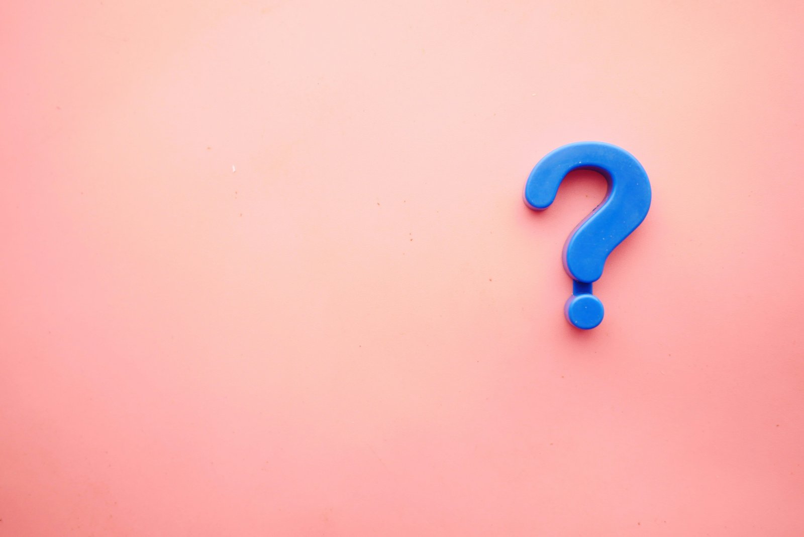 A blue plastic question mark resting on a pink background.