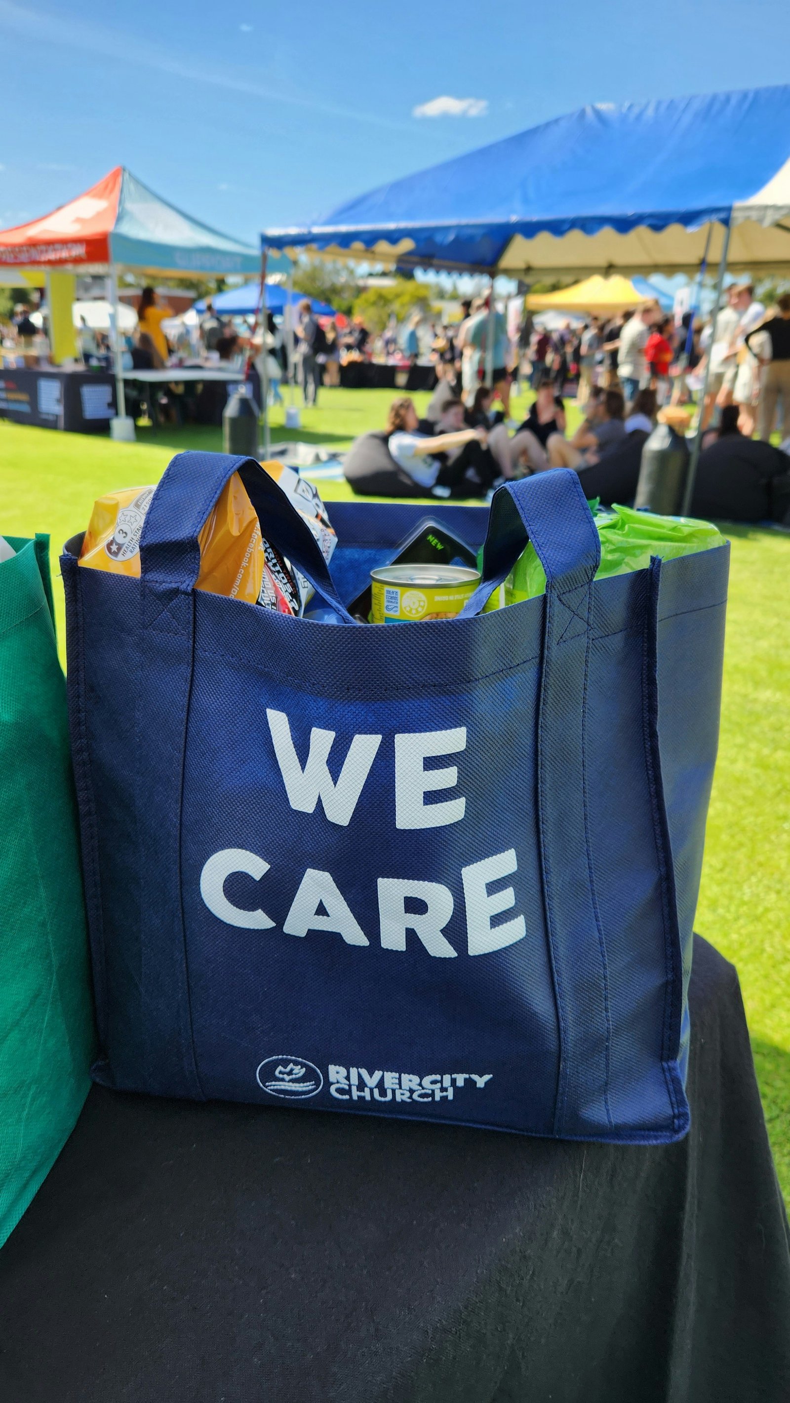 A 'We Care' tote bag filled with groceries at an outdoor community food drive.