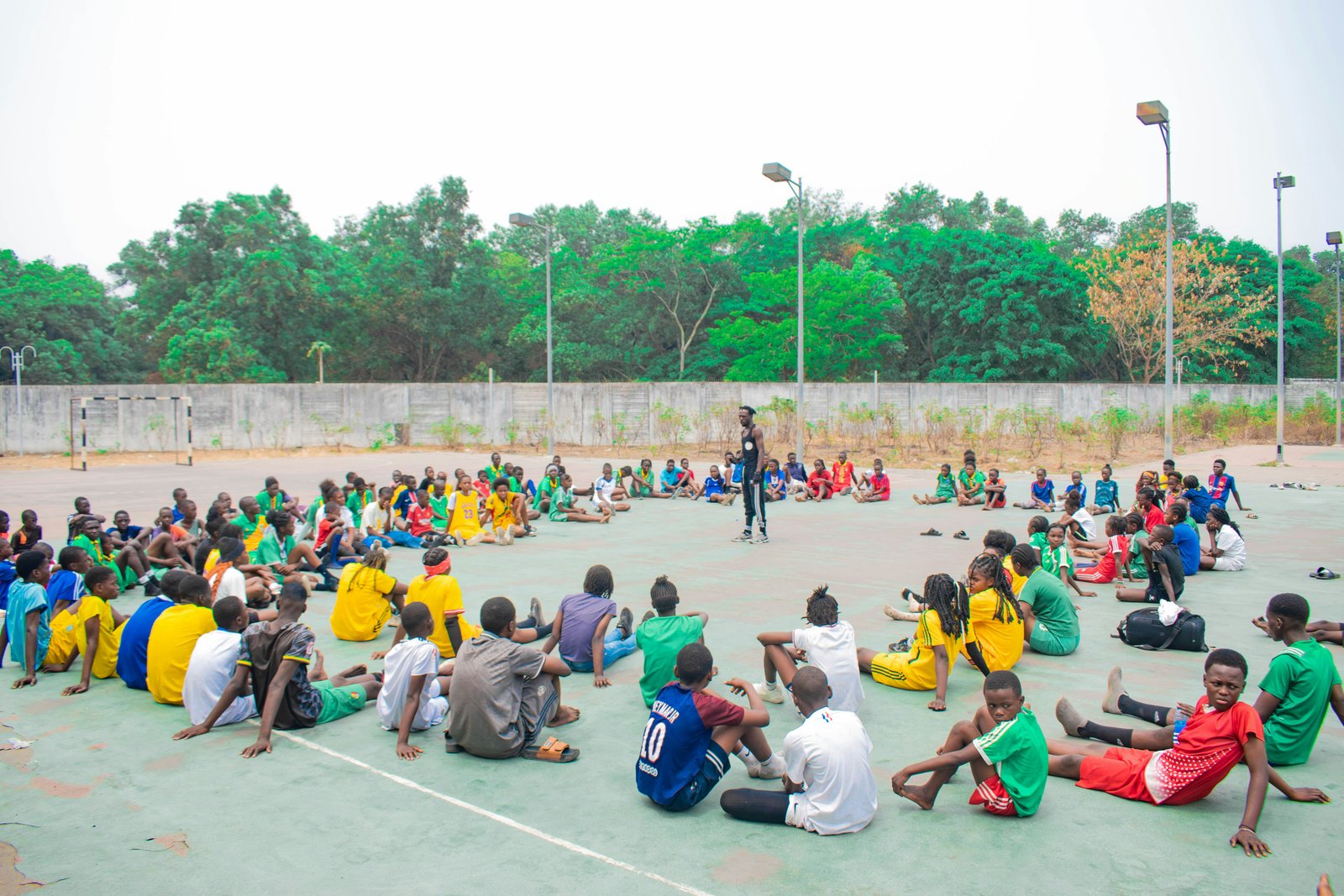 A large group of youth sitting together outdoors listening to a coach.